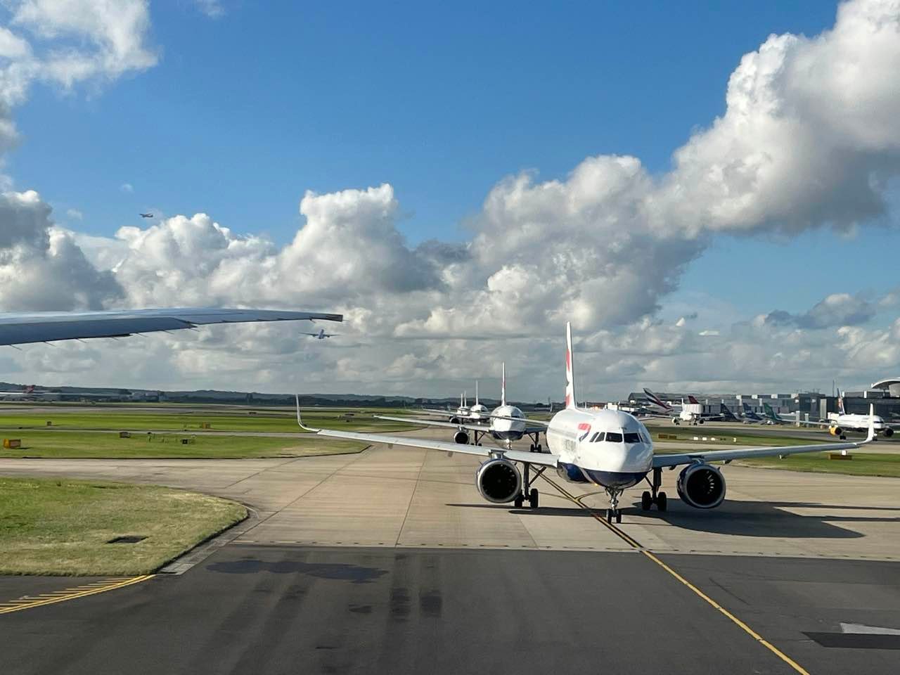 British Airways planes queuing on a taxiway at Heathrow Airport under a blue sky with white clouds
