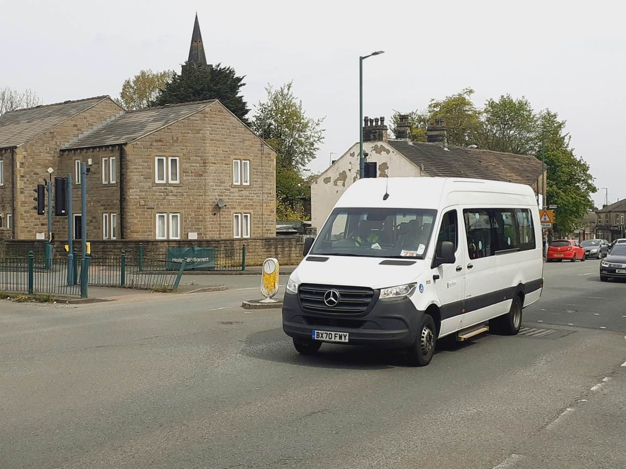 A white minibus driving along a road past stone houses in Bradford, West Yorkshire