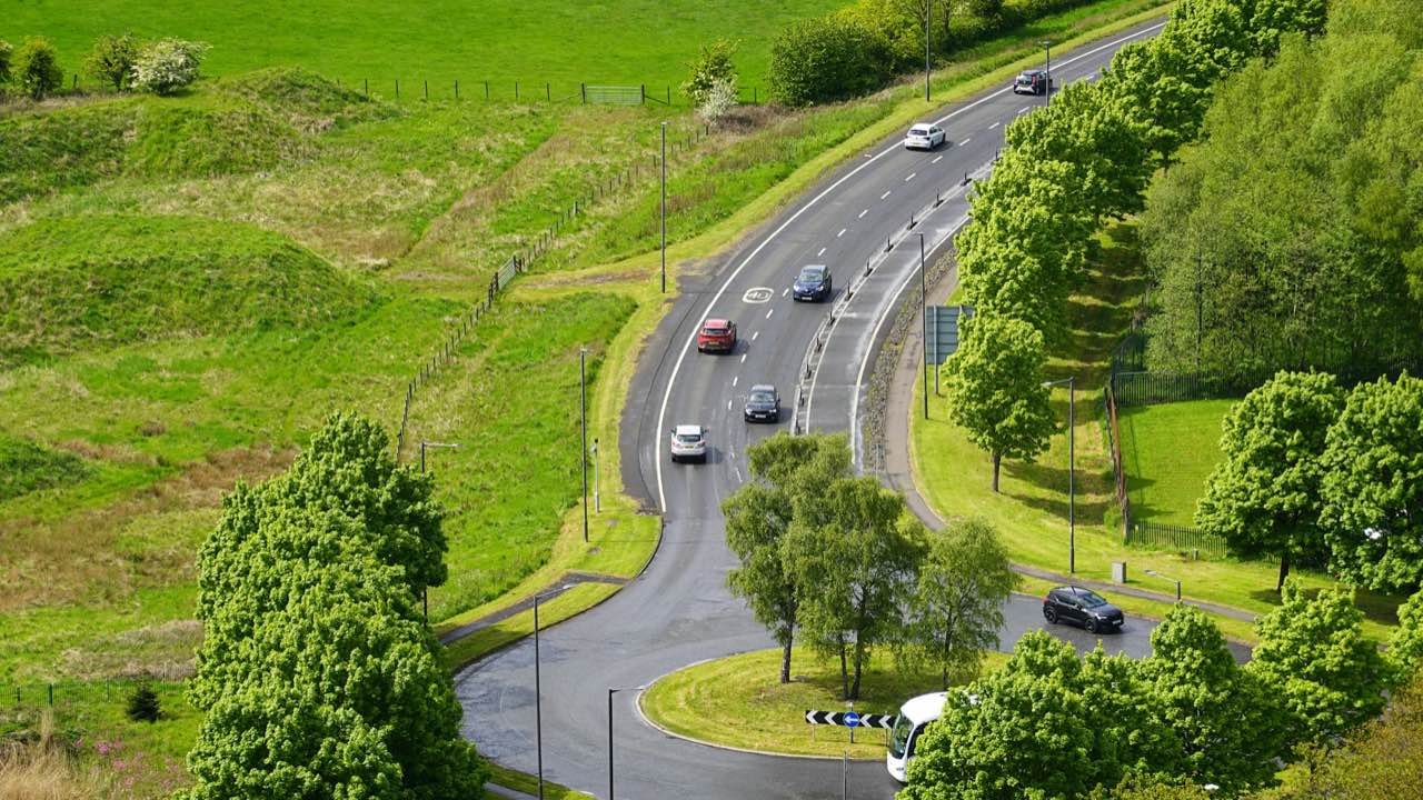 A road curving down to a roundabout between green fields and trees