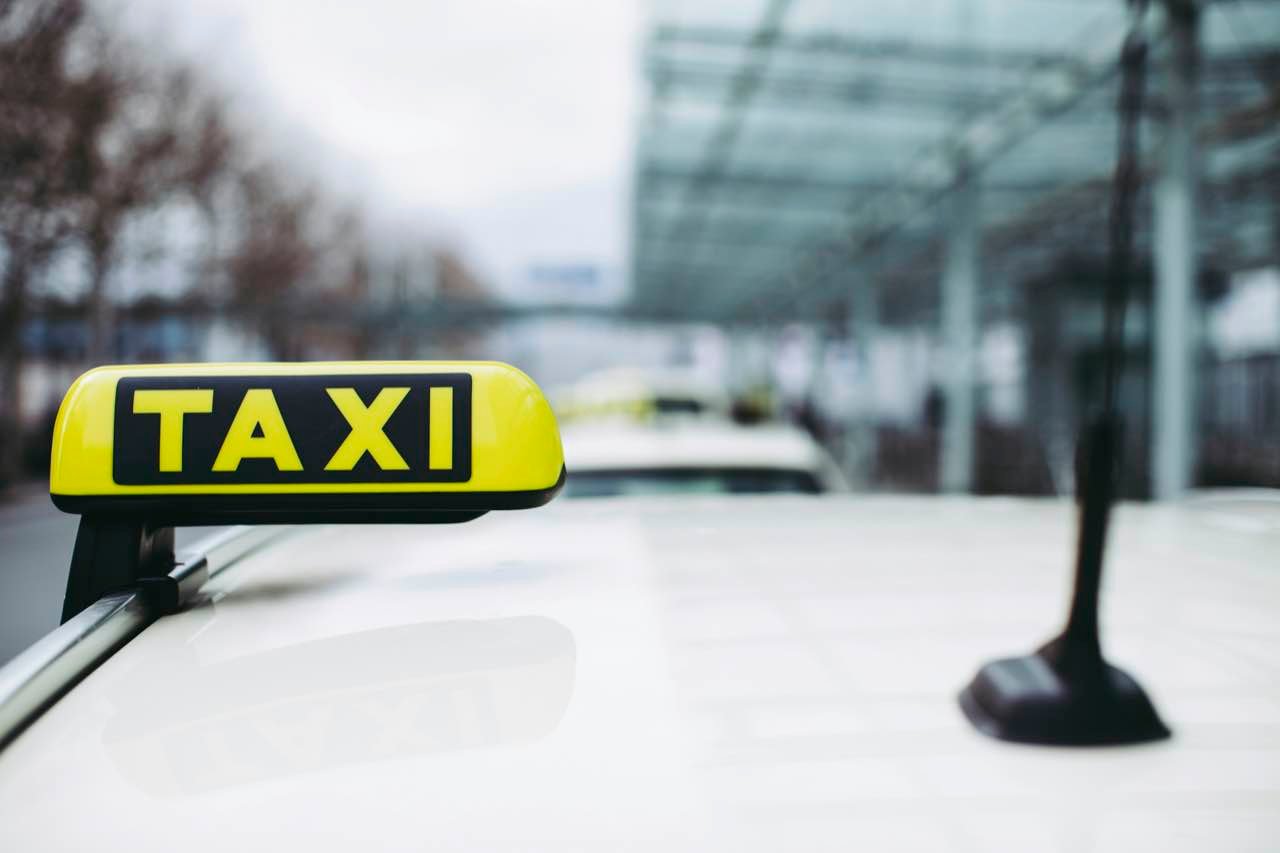A yellow TAXI sign on the roof of a white car outside a glass-roofed building