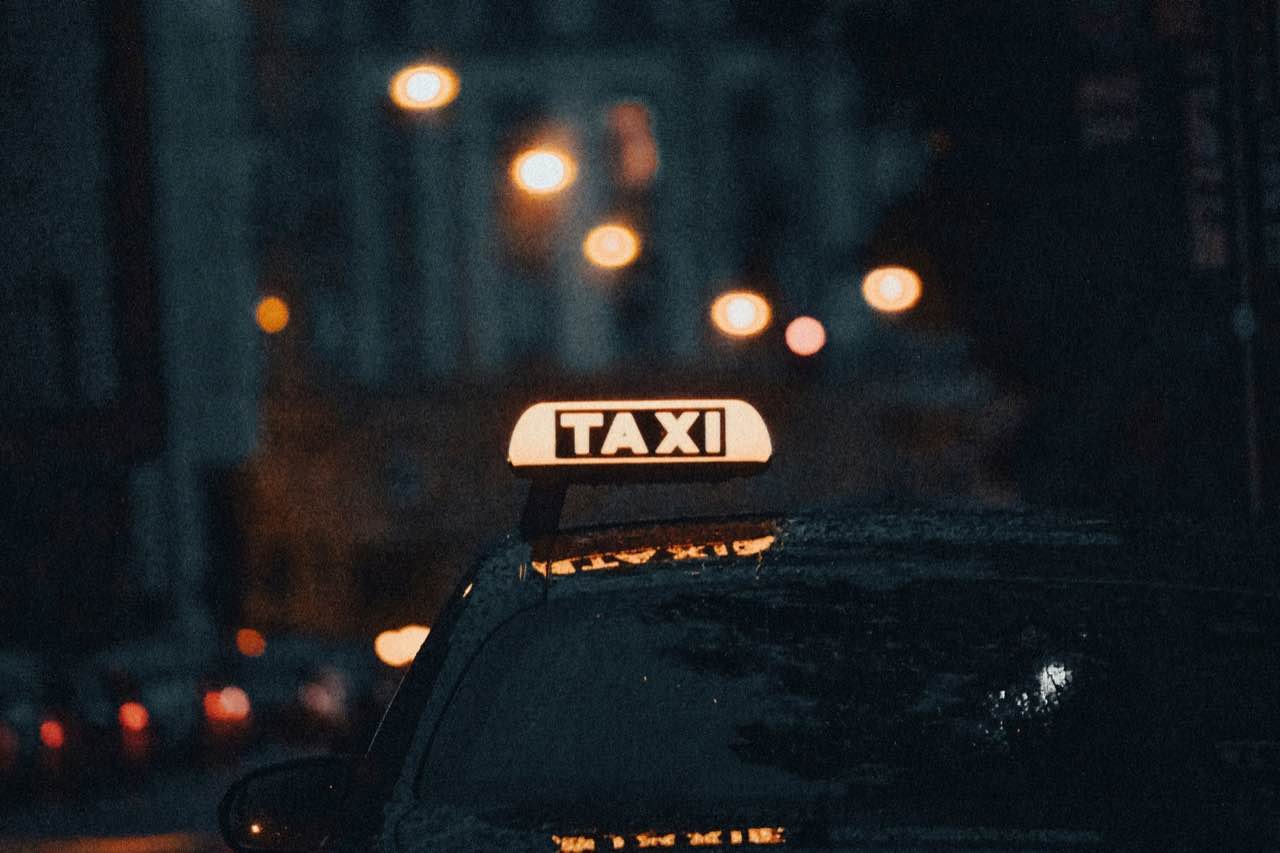 A lit white TAXI sign on a dark car at night, with street lights blurred behind
