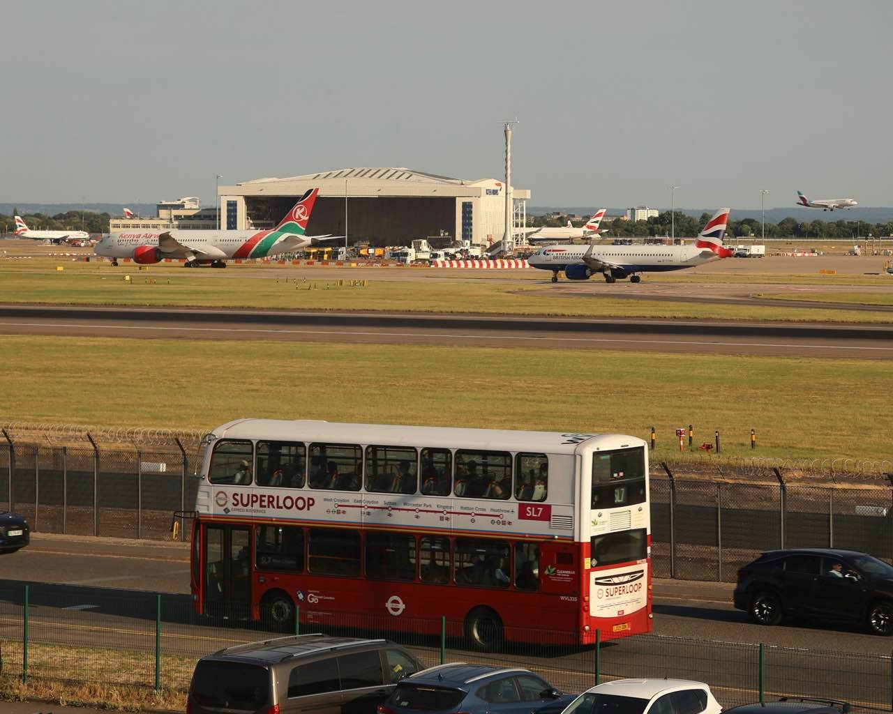 A red Superloop double-decker bus on the road beside Heathrow Airport, with planes on the airfield behind